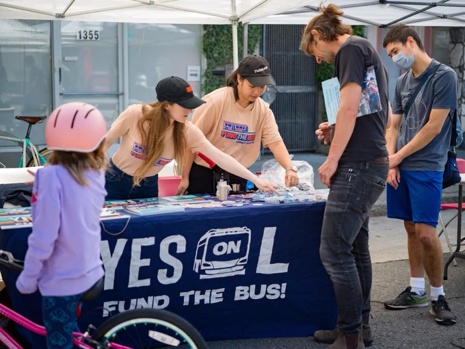 Two volunteers behind a table that says "YES ON L FUND THE BUS"