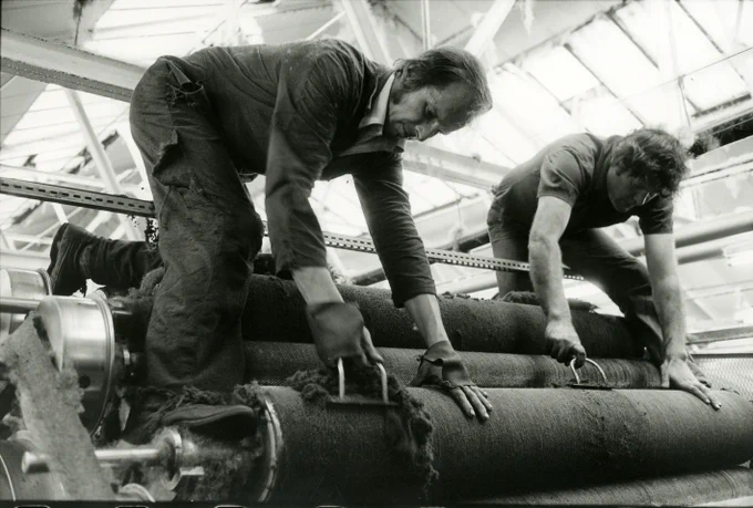 A black and white photograph showing 2 men hard at work atop a large machine with two rollers. It's called 'Card Fettlers, Brackendale Mills, Thackley, Bradford, 1980s' and it's by Ian Beesley. Of the photo, Beeslet comments: “With the demise of traditional industry many of the local dialect words peculiar to specific trades and industries have vanished from our vocabularies: the jobs are extinct and so are the words to describe them. I find that deeply sad. I’ve always loved these job titles and hope my work will in some small way stand testament to the folks who grafted through their lives doing these jobs."
