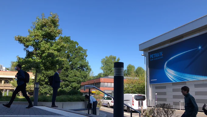 Menschen gehen unter einem Banner mit der Aufschrift "PETRA IV" unter einem blauen Himmel und zwischen Bäumen hindurch
