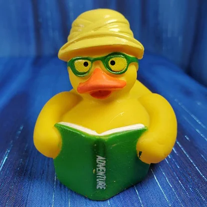 A small yellow rubber duck wearing a safari hat and green-rimmed glasses is reading a green book with the word "Adventure" showing on the spine.