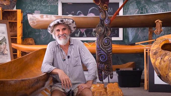 Person sitting between two Polynesian canoes in a room with cultural artifacts, surrounded by chalkboards covered in writing and diagrams.