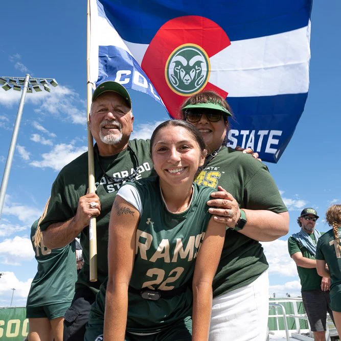 C S U soccer player Mia Casey posing for a picture with two people decked out in Rams gear and holding a large C S U flag.