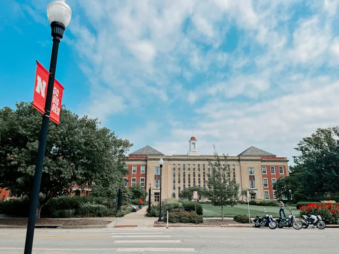 Love Library on a sunny day. A banner on a light post reads "Go Big Red" and people in the distance walk past the library and work in the garden.