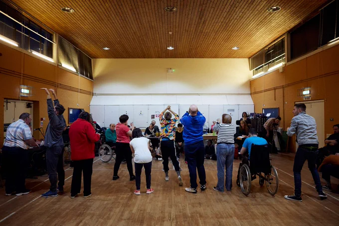 Lung Ha actors taking part in a workshop. They are lined up in a row and striking various poses. They are in a wood-panelled room with a white wall in the background of the image.