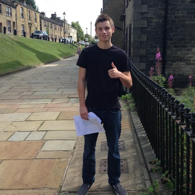 Callum stood on the front terrace of his high school, Fulneck School, with the Church in the background. He is holding up the document with his GCSE results.