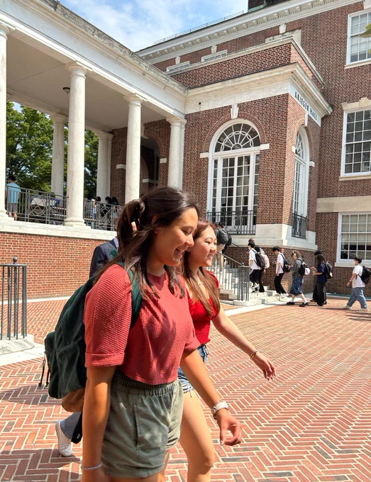 Two smiling students are walking on the Homewood campus surrounded by other students and large brick buildings in the background.