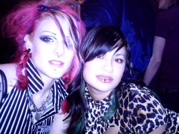 Photo: Two women sat posing together. The woman on the left has bright pink hair, heavy eye makeup, a lip piercing, and is wearing a black and white striped top. The woman on the right has long black hair with green highlights, a lip and septum piercing, and is dressed in a leopard print top. 