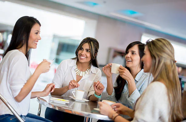 4 women sit around a table, smiling and drinking coffee. 
