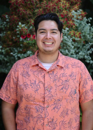 Jasper Gomez smiles at the camera. He is wearing a coral-colored shirt with blue octopuses all over it and standing in front of colorful plants. 
