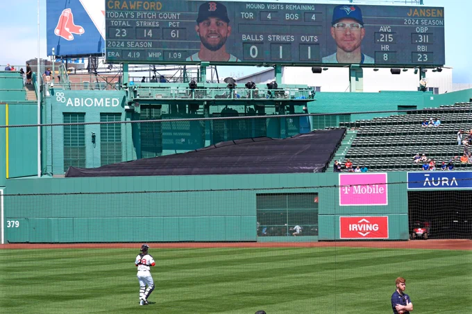 Danny Jansen stands in the outfield of Fenway Park in a home Red Sox uniform. In the background is a headshot on the videoboard of Danny Jansen in a Blue Jays uniform.