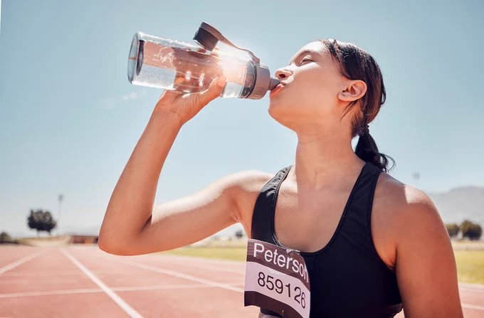 A runner drinks from a clear sports bottle as she stands on a track.
