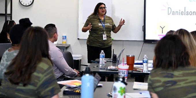 Person presenting at a workshop in a classroom setting, standing in front of a screen displaying the name "Danielson." Participants seated around tables, listening and taking notes.