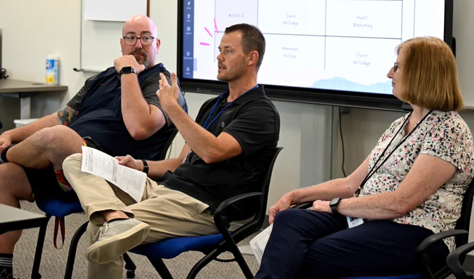 Three teachers are seated during a panel discussion in a room with a projector screen displaying a presentation in the background.