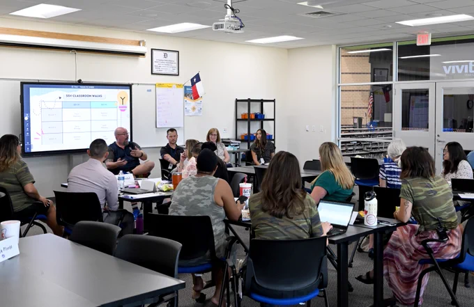 Adults seated at desks in a classroom setting, attentively participating in a presentation led by a facilitator pointing at a screen displaying a bar graph and text. The room has educational posters and flags, illustrating a collaborative learning environment.