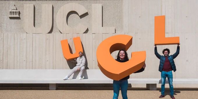 Three people pose with large orange letters spelling "UCL" in front of a concrete building at University College London.
