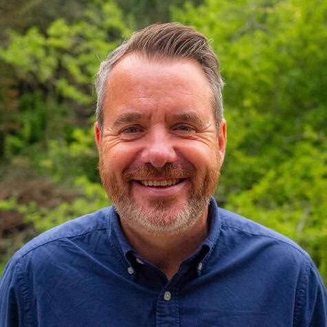A Caucasian man in a blue shirt smiling. He has brown hair and he is outside. There is greenery behind him.