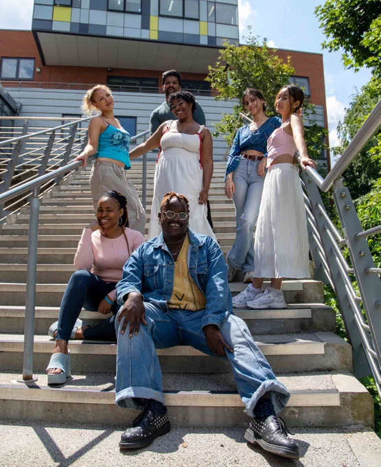 A group of six students posing on a staircase outdoors, smiling on a sunny day. They are dressed in casual summer attire.