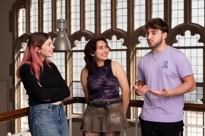 Three University of Manchester students stand in Manchester Museum talking to each other. The boy on the right hand side is talking and using his hands to gesture, while the two girls on his left look at him and smile.