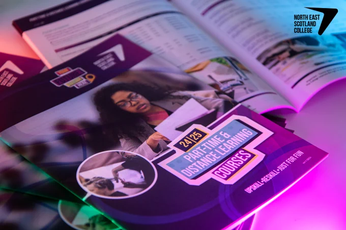 Guides for the 2024/25 part-time and distance learning courses at North East Scotland College, featuring an image of a focused student reading a document. The guidebooks are arranged on a table with a vibrant purple and blue lighting effect highlighting the materials.