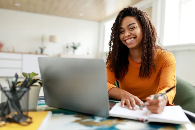 A person smiling at a laptop computer while writing in a notebook.
