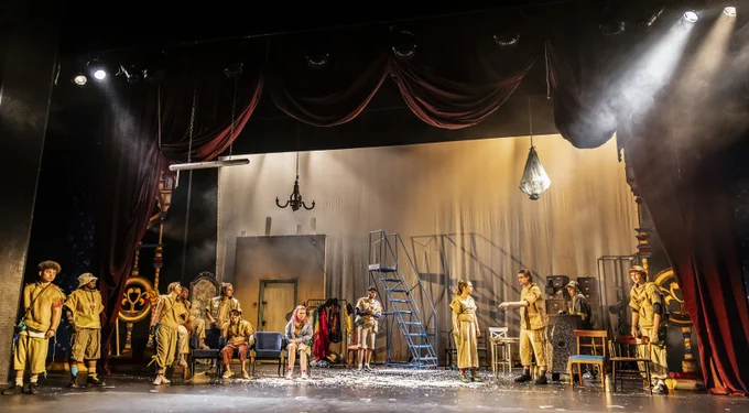 A stage filled with props, sets, and costumes from past shows. Twelve young people, wearing clothes made from hessian sacks and coffee bags, populate the space. Some are sitting on chairs, but most are standing. They are all looking towards one of the young people, who is pointing directly in front of them.