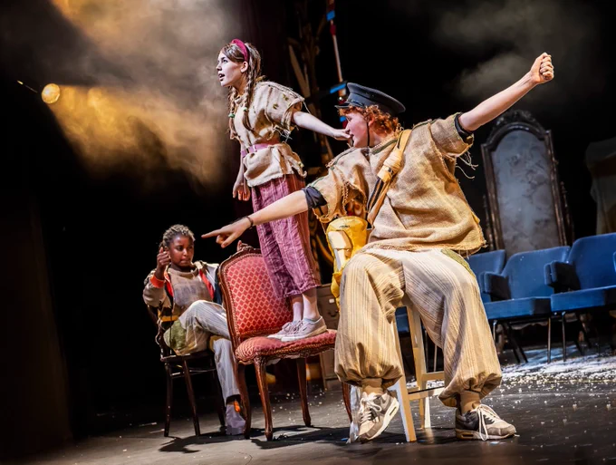 Three young people on a disused theatre stage wear clothes made from hessian sacks and coffee bags and sit on chairs one behind the other. They have their arms out wide and look excited. A stage spotlight lights the stage.
