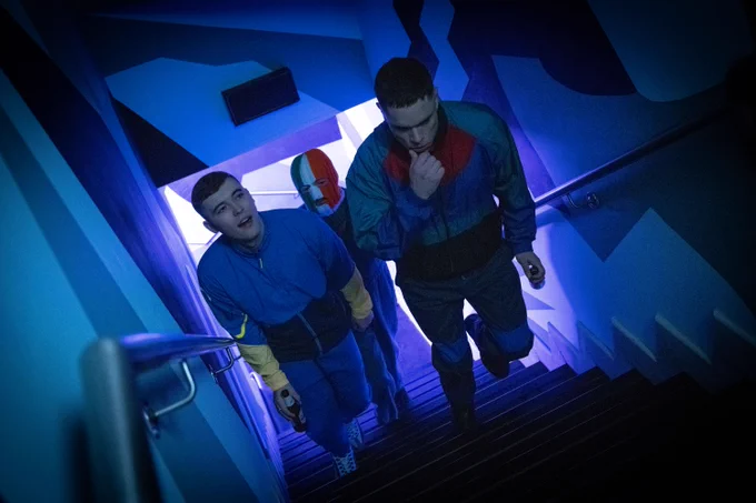 Still from Kneecap film showing the three band members walking up the stairs in what looks like a dimly lit club. Two of them are carrying beers and one of them is wearing an Irish flag balaclava.