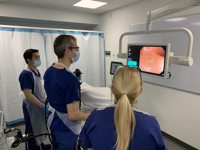 Three healthcare professionals, two standing and one seated, are focused on a monitor displaying medical imaging during a procedure in a hospital room.