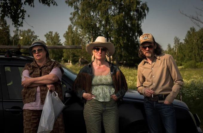 A film still from 100 Litres of Gold. Three people are standing against the side of a car, all wearing sunglasses and hats and holding stern expressions, as a field and trees fill the background. 