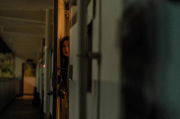 A still from Noise. A women is peering through a slightly ajar door with a frightened expression, staring at a figure that's out of focus in the foreground.