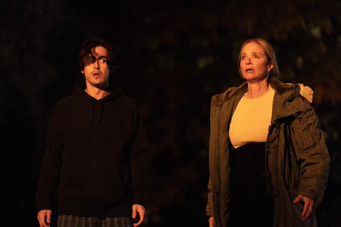 A still from Yuppie. A man, wearing a black hoodie, and a woman, wearing a light top and dark pants with a large jacket, are standing outside in the dark, yet bathed in a bright orange light, both with shocked and frightened expressions.