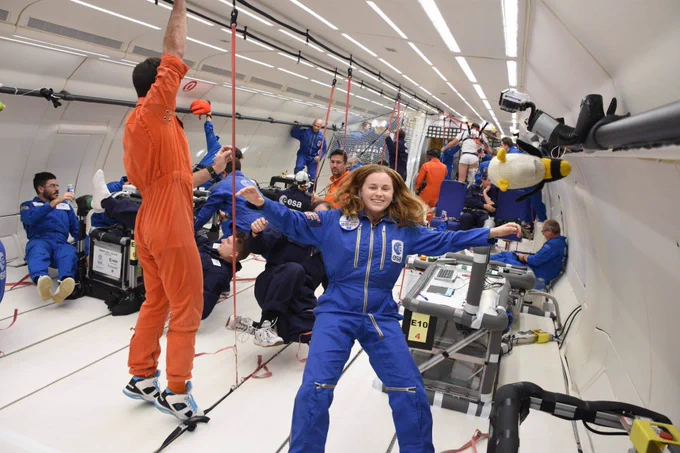 A woman in a blue ESA boilersuit floats inside a parabolic flight plane, with a number of other trainee astronauts in similar outfits behind her