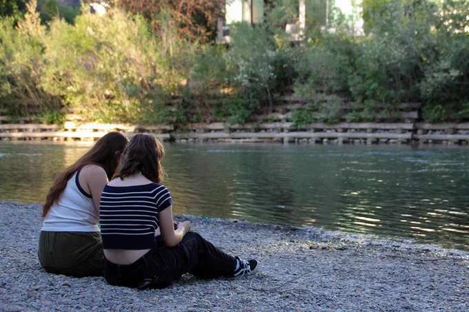 Two campers sit quietly together on a riverbank, facing the green water. 