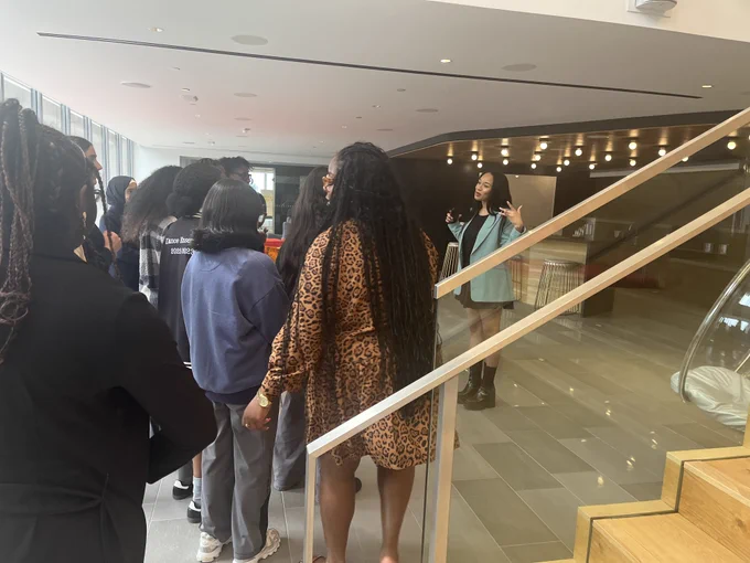 Students stand in a group and listen to a woman speaking to them. 