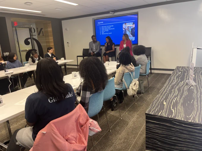 Students sit at a long table in a room listening to panelists at the front. 