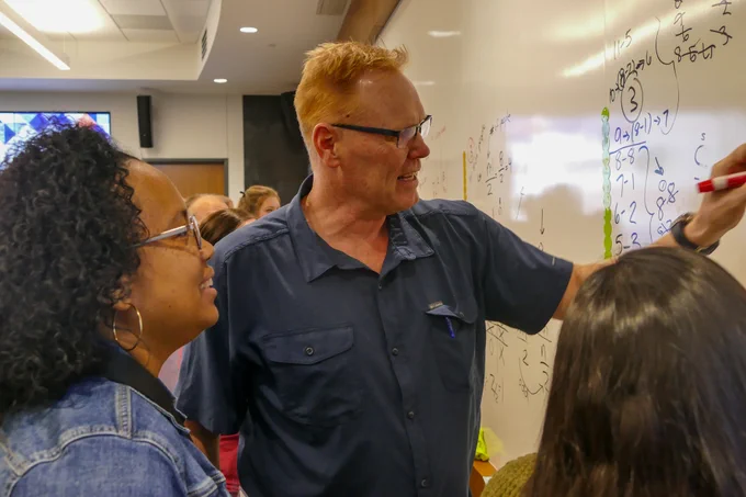 Three people engaging in a discussion while Peter writes mathematical equations on a whiteboard.