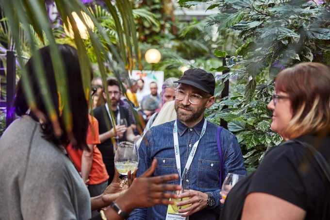 A group of people are engaged in conversation at an indoor gathering surrounded by lush green plants. A person wearing glasses and a hat is holding a drink and smiling, while others around also hold beverages. The atmosphere appears casual and social.