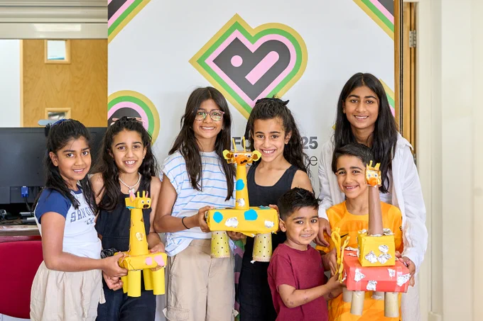 A group of children smile into the camera holding model giraffes made from craft items. In the background is a brightly coloured logo in the shape of a heart. 