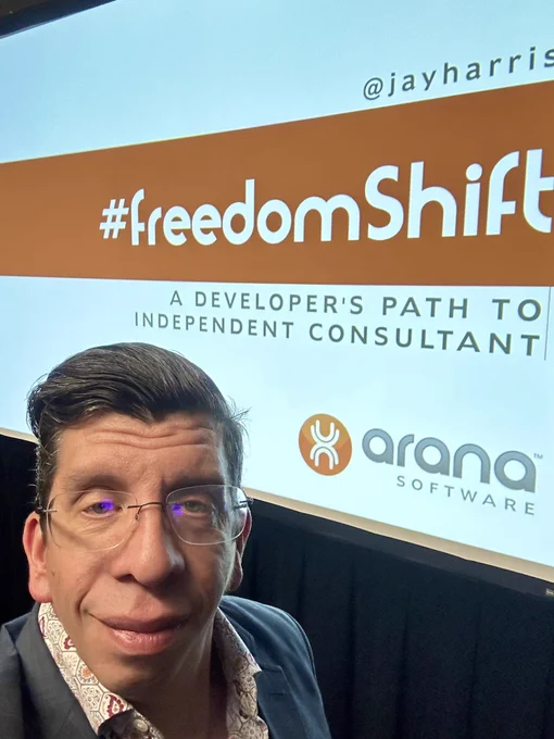 A selfie of Jay Harris in a gray-blue suit coat standing in front of the projected image of the title slide to “Freedom Shift: A Developer’s Path to Independent Consultant.”