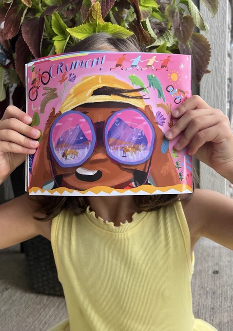 Little girl holding up a page of Armando's book "My Great Adventure with Nana" so that an illustration of a child's face is covering her own face.