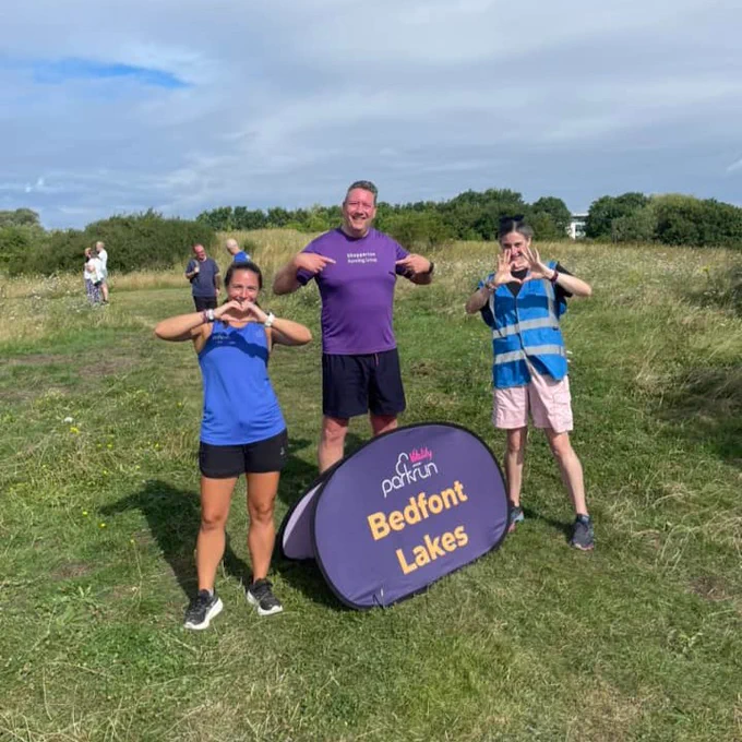 Three parkrunners pose at Bedfont Lakes parkrun, with two making heart shapes with their hands. 
