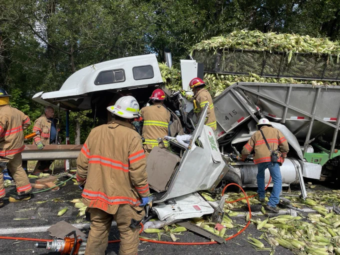 Firefighters examine the damage and work to pull the injured driver from the cab of the truck on the freeway. Spilled corn litter the roadway.