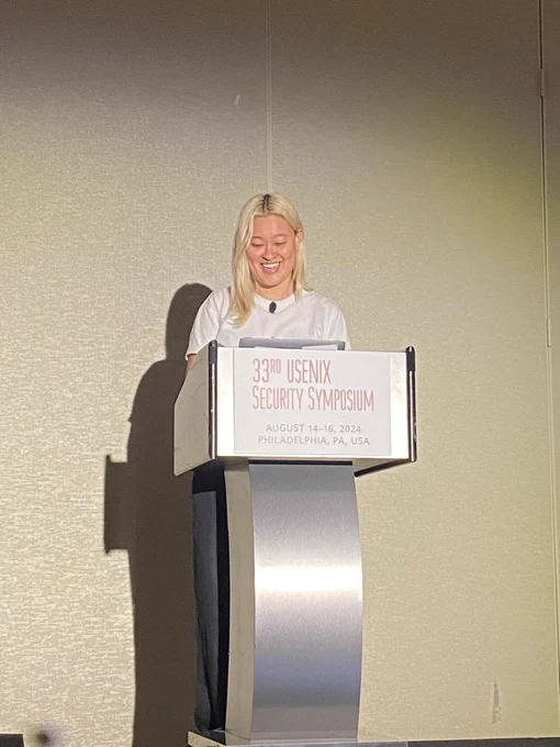 Miranda, a blonde Asian person, stands and laughs from behind a lectern that has a sign for "33rd USENIX Security Symposium"