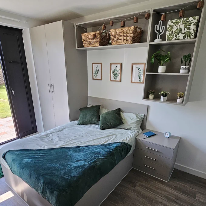 A bedroom. Green blankets and cushions are on a double bed. Large open shelves above the bed hold baskets, plants and decorations. The room is on the ground floor and a grassy space can be seen outside the large window.