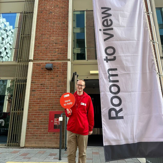 An ambassador in red hold up a paddle that says "here to help". He is stood in the doorway of an accommodation block. A big white vertical banner next to him reads "Room to view"