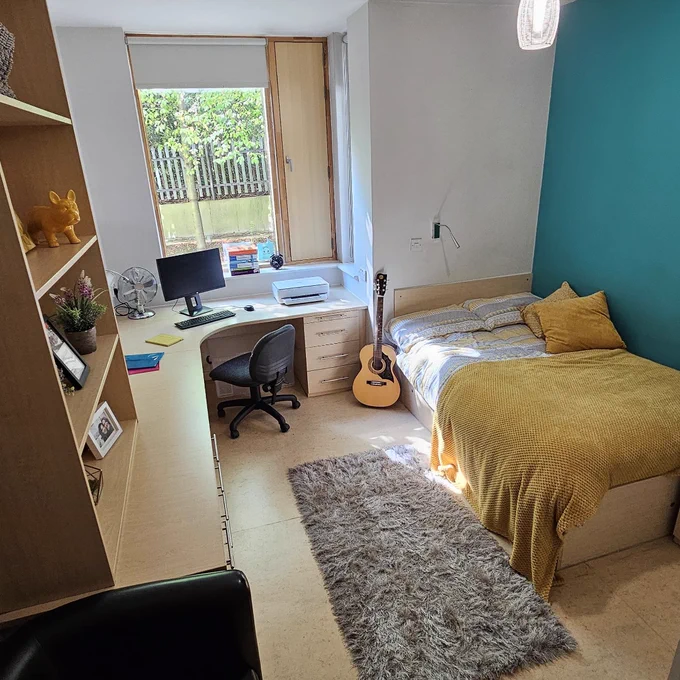 A bedroom. The room has a large curved desk and shelves with a computer and personal belongings. A large bed is in the corner decorated with a yellow blanket. An acoustic guitar rests against the bed.