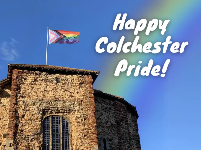 A Progress Pride flag waves atop a historic stone building with a large arched window. The building has a reddish-brown roof and is set against a clear blue sky. A rainbow effect is overlaid on the right side of the image, behind bold white text that reads "Happy Colchester Pride!"