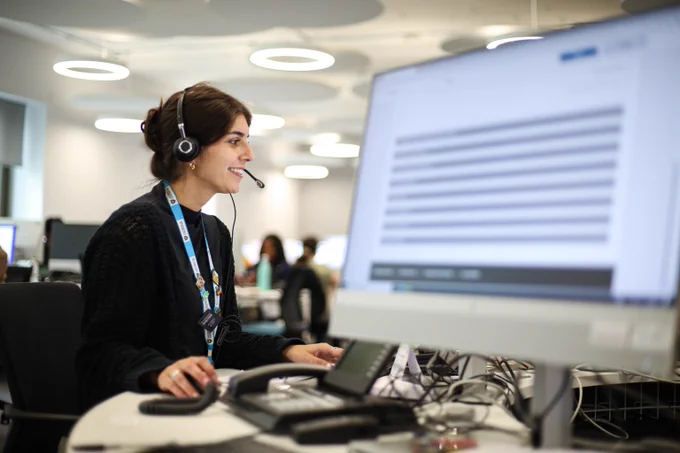 One of our friendly call handers is sat at a table talking on a headset. In the foreground is a computer screen and in the background other call handlers can be seen at their desks.