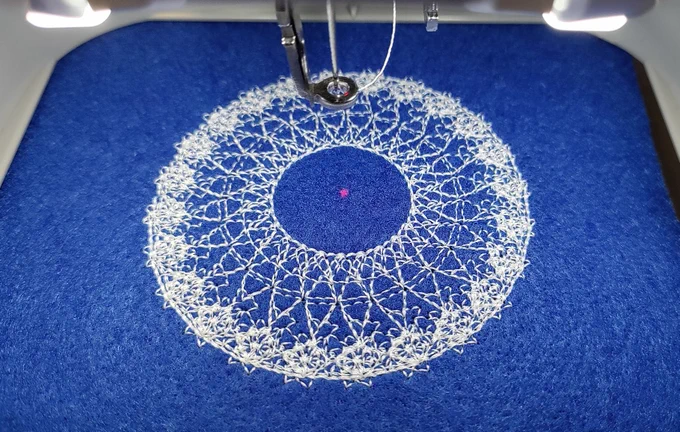 Circular stitched pattern that looks like a flower, stitched with white thread on a blue felt background. 