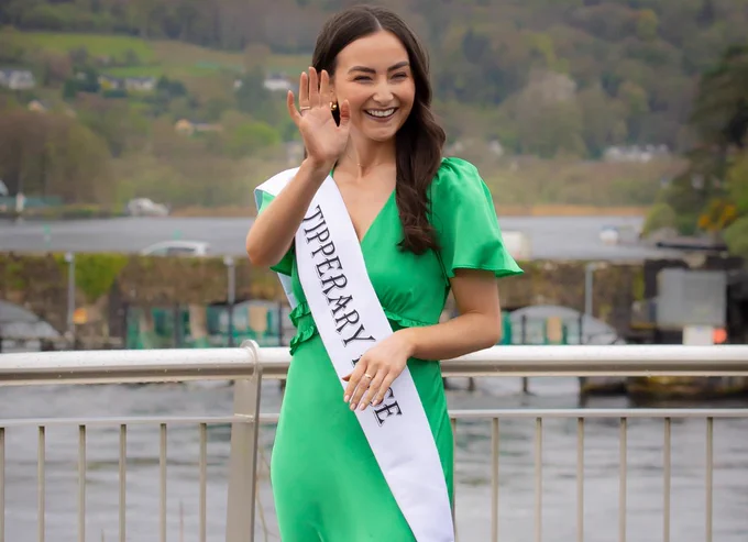 A photograph of a woman in a bright green dress. She is wearing a white sash with the words "Tipperary Rose" written on it. She is seen on a bridge over a wide river and is waving at the camera with her right hand